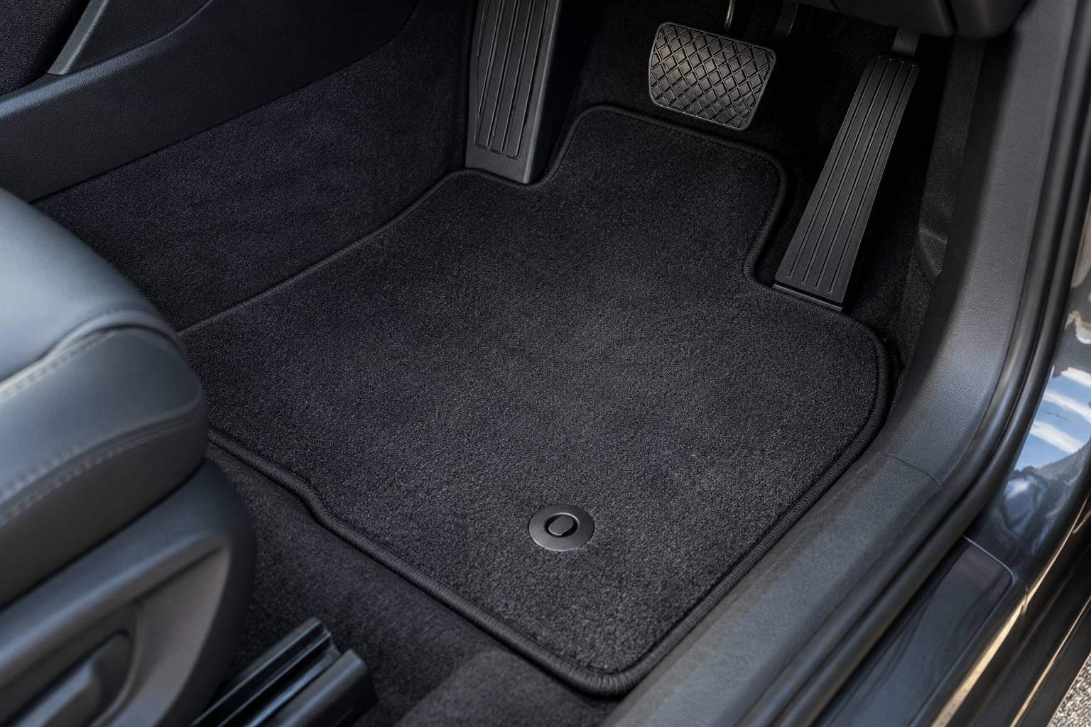 A tailored charcoal carpet mat fitted in a right-hand-drive footwell, its cut edge following the tunnel and sill, with a fixing stud engaged through the mat