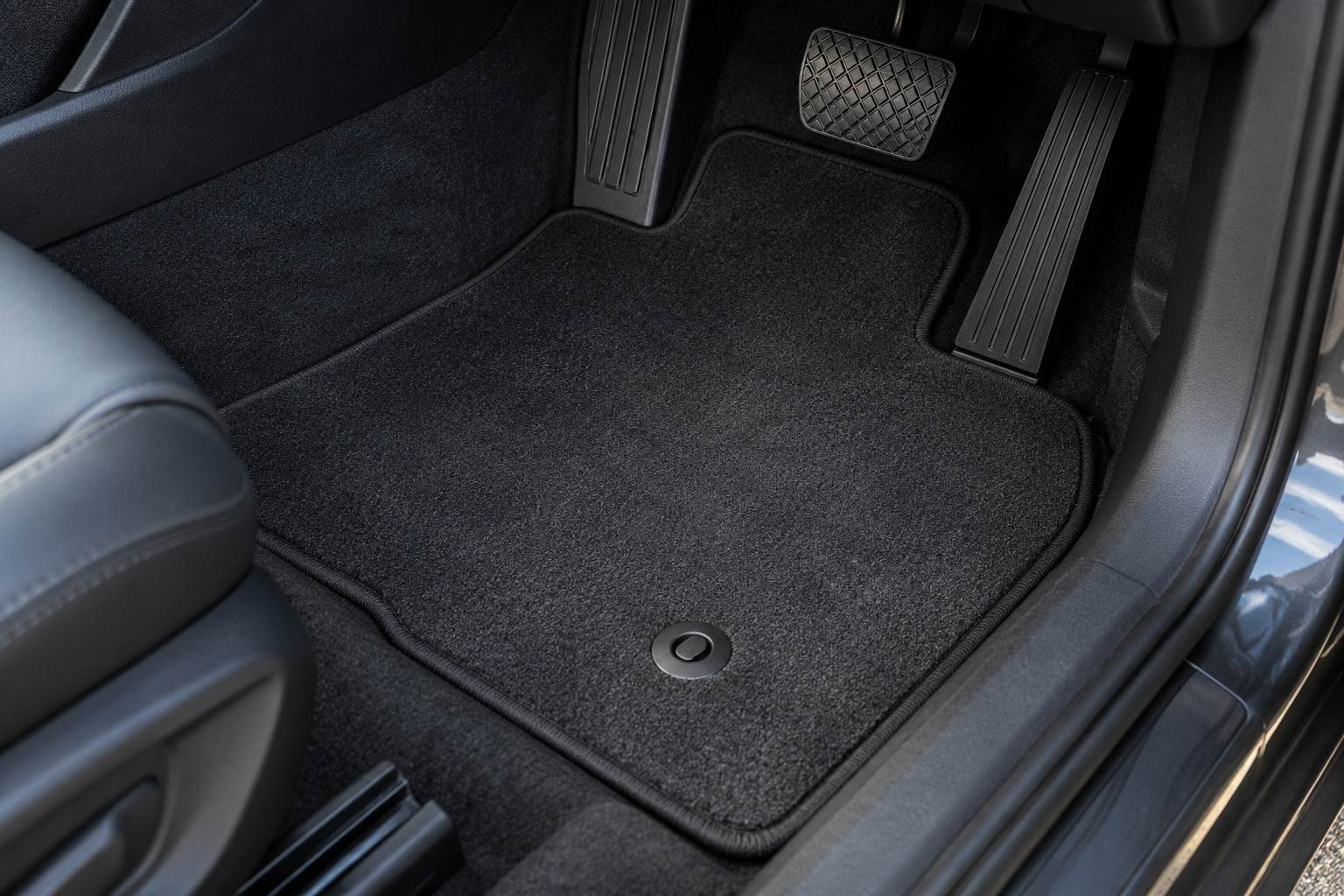 Tailored charcoal carpet mat fitted in a right-hand-drive footwell, its cut edge following the tunnel and sill, with a round fixing stud engaged through the mat